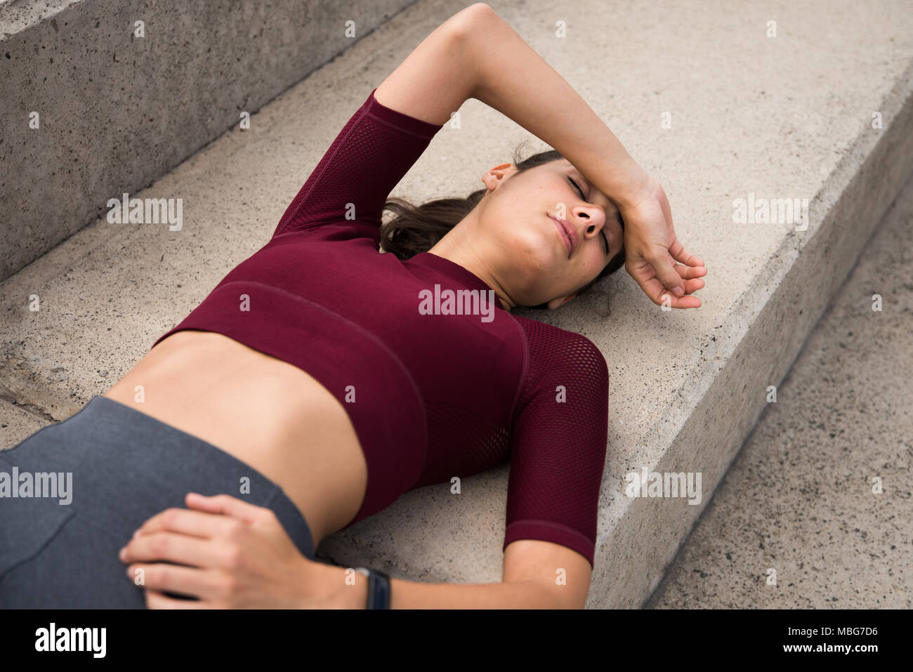 Sporty young weary brunette resting on stairs after work out wearing grey tights and maroon midriff shirt Stock Photo