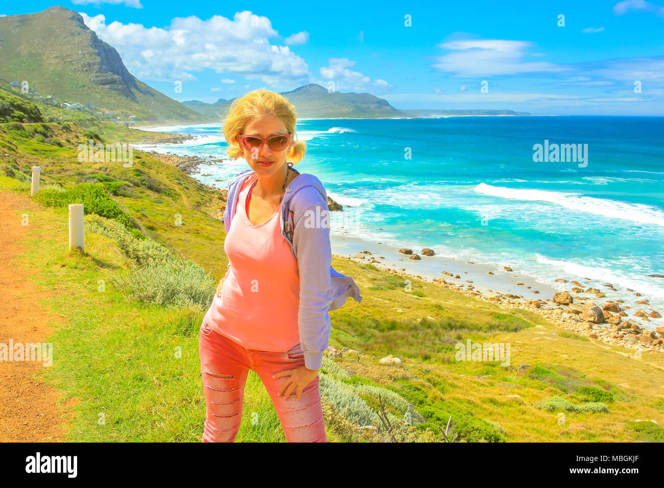 Blonde female tourist on popular attraction Chapmans Peak Drive in Cape Peninsula near Cape Town, South Africa. Lifestyle woman traveler in day trip road. Summer holidays. Blue sky. Stock Photo