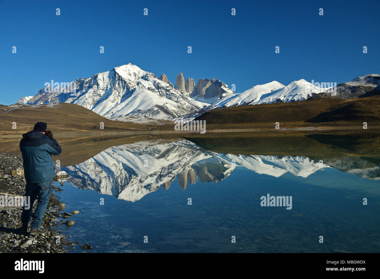 Lone tourist standing on the shore of Laguna Amarga with the Torres del Paine massif reflected in the mirror calm waters. Stock Photo