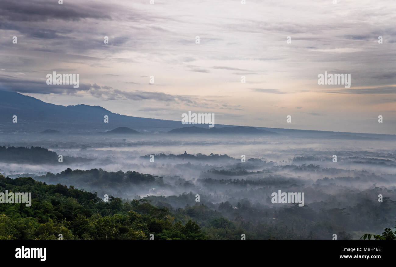 view of the Kedu Plain in the early morning mist with the distant stupas of ancient Borubudur Buddhist temple rising above the plain, seen from Punthu Stock Photo