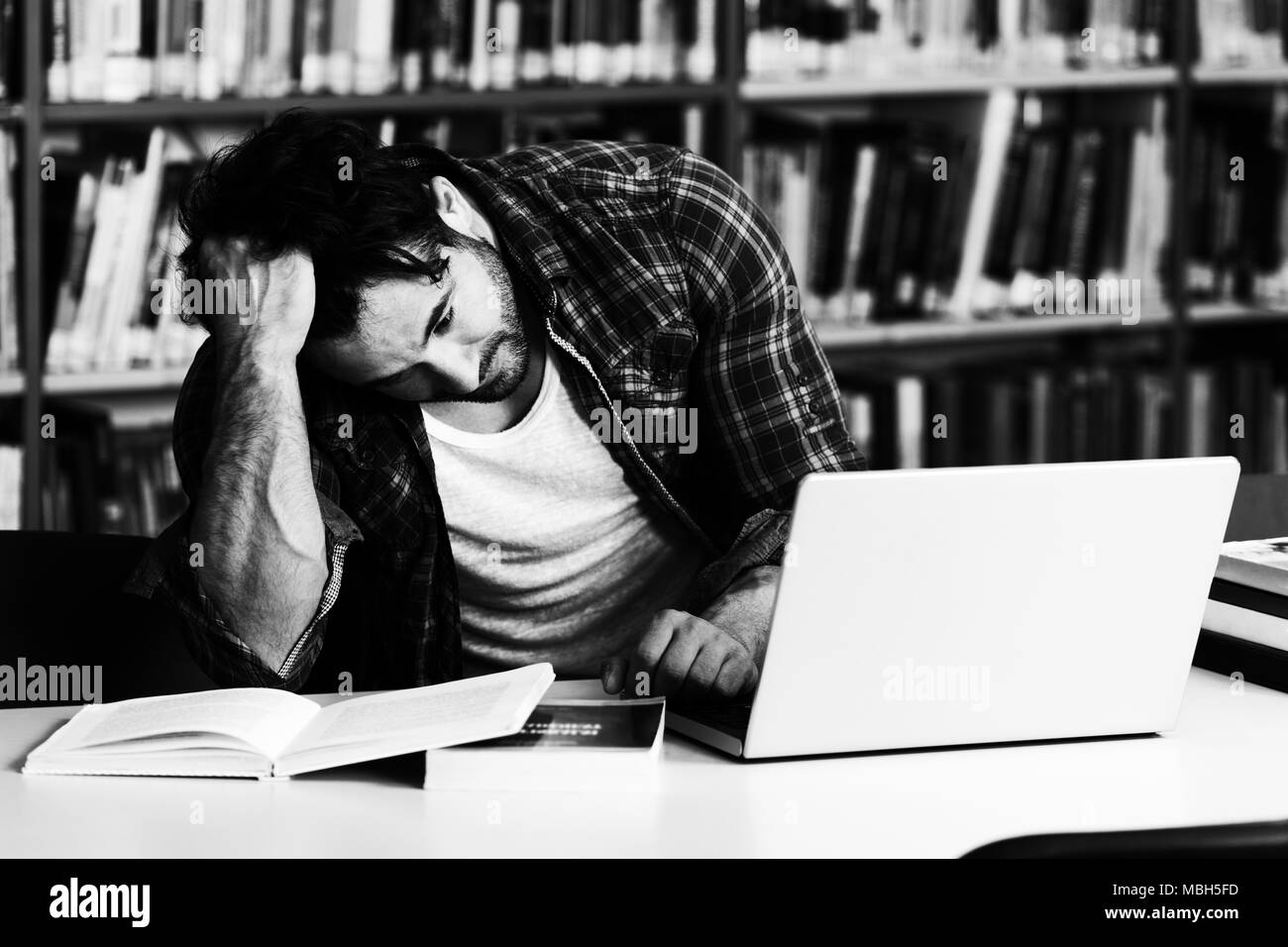 Male College Student Looks Tired While Studying With a Laptop and Textbooks in the Library Stock Photo
