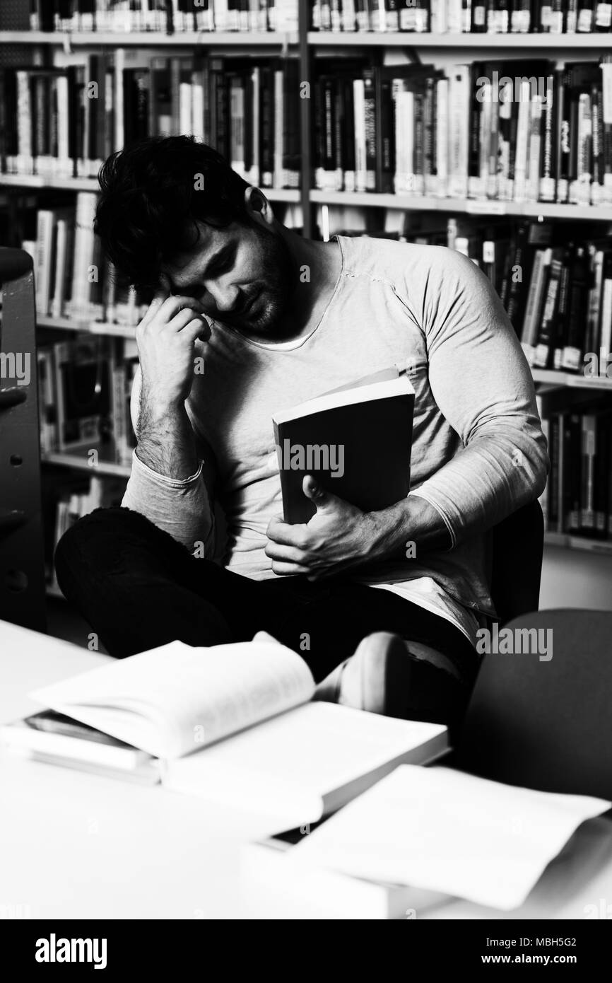 Stressed Young Male Student Reading Textbook While Sitting in Library Stock Photo