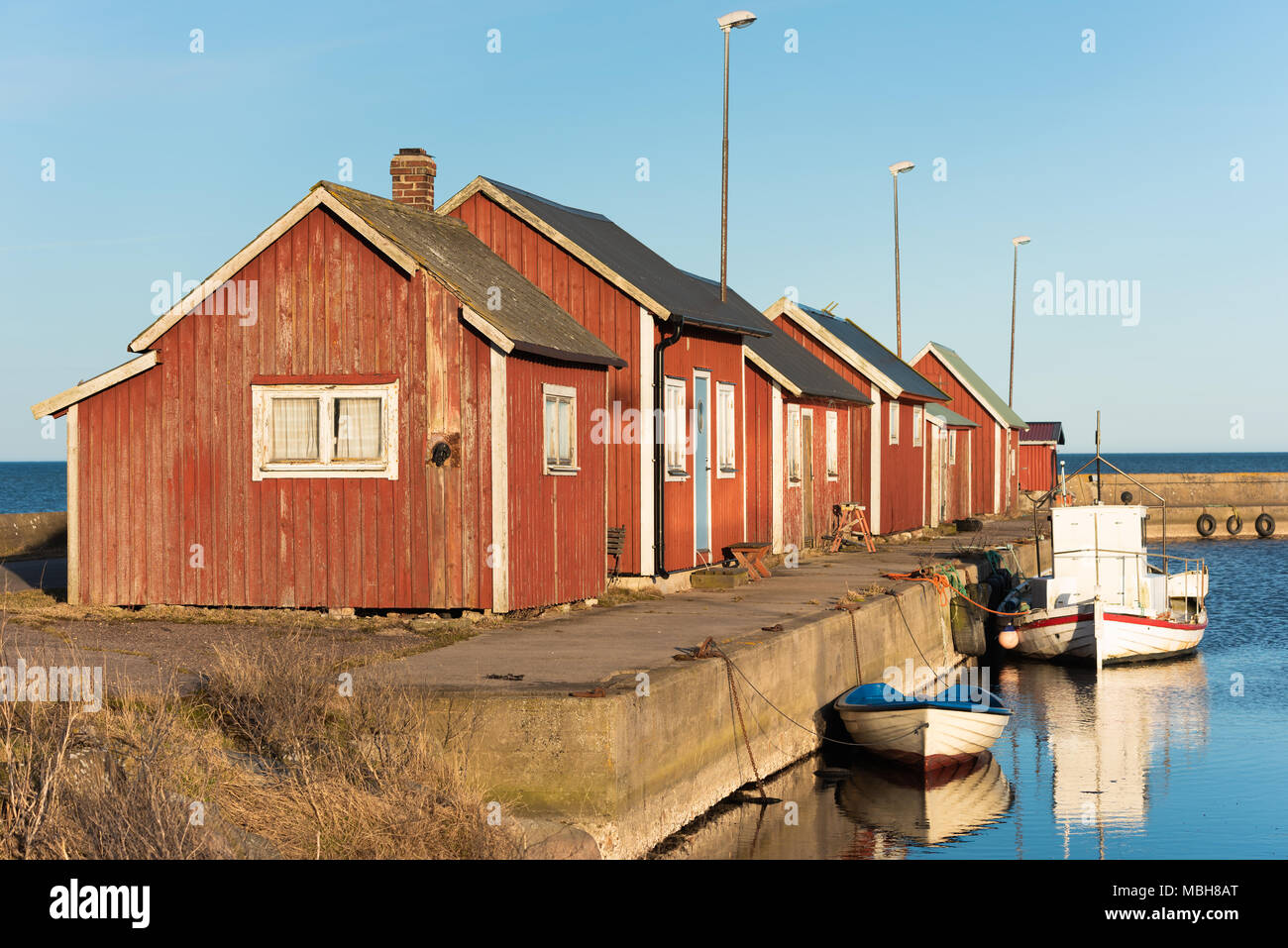 Gammalsby harbor at eastern Oland, Sweden. Small red wooden fishing cabins in a row on the pier with small boats moored dockside in the evening sunlig Stock Photo