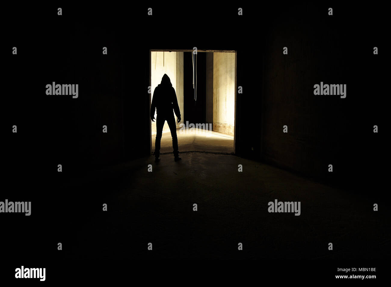 unrecognizable silhouette of person with a hood standing in a door way of an empty concrete room. Stock Photo