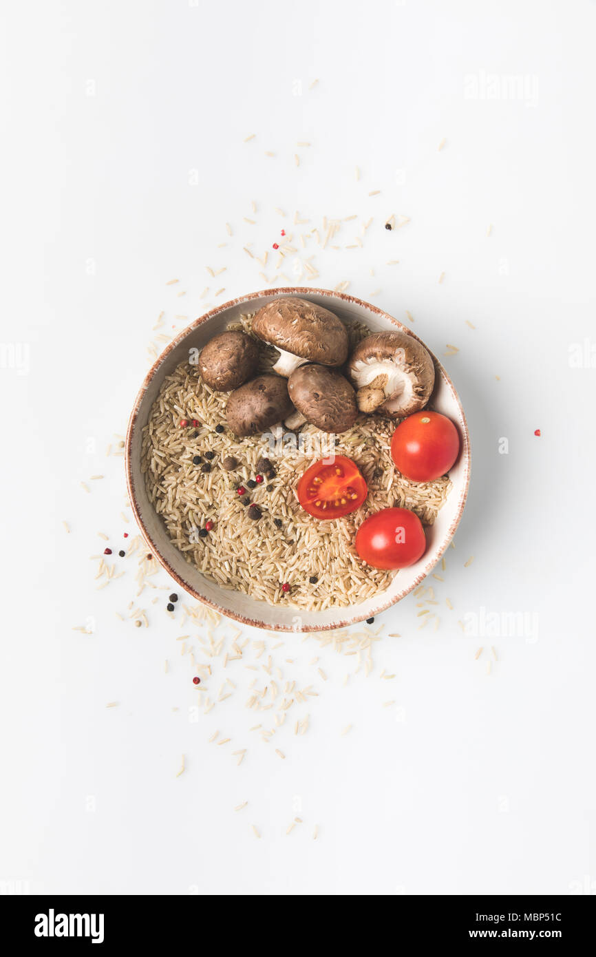 top view of raw rice in bowl with mushrooms and tomatoes on white ...