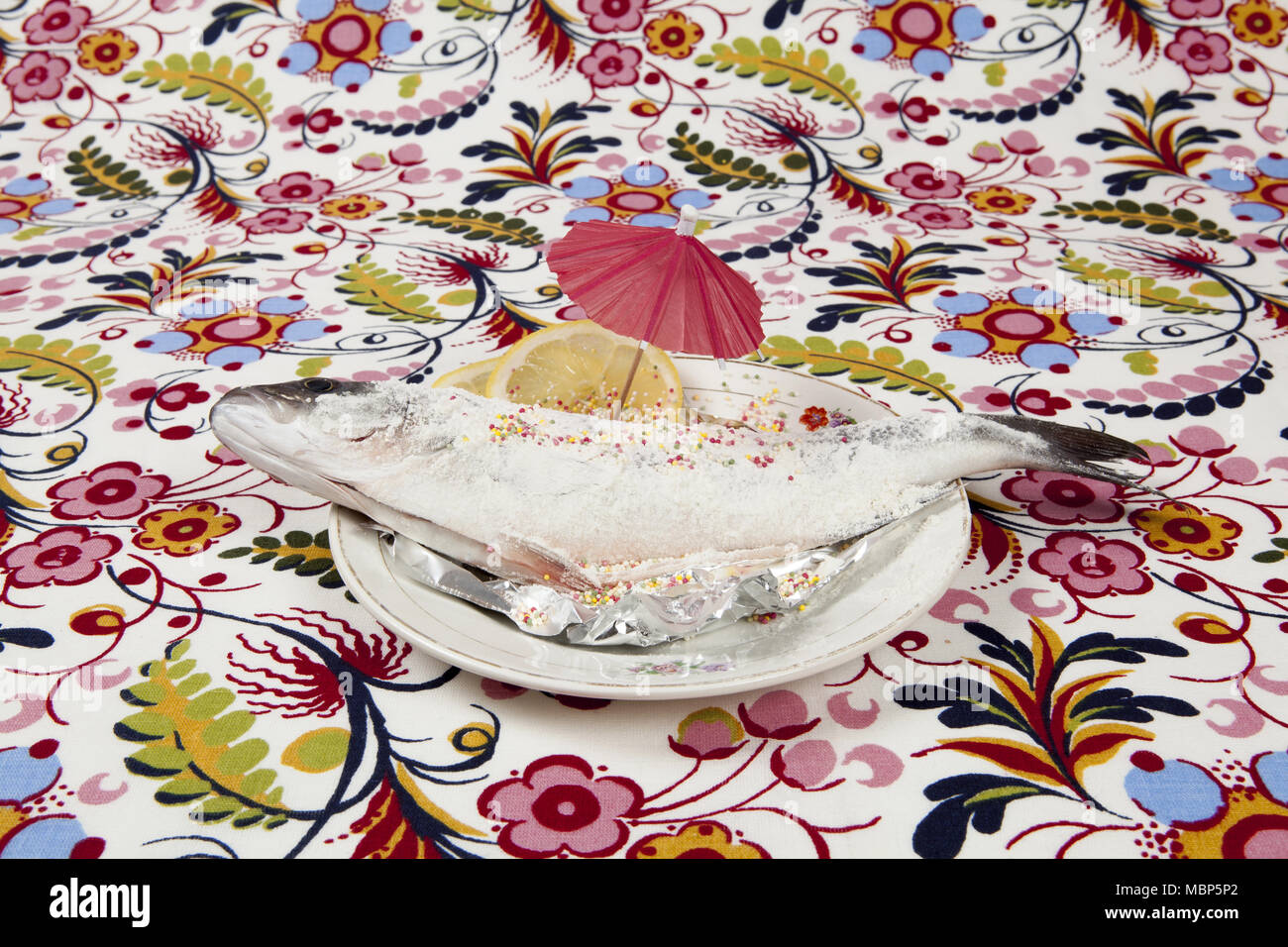 A preparation of a bass fish with sprinkles and a slice of lemon beside inside a flower plate hidden on a flowery fabric. Camouflage game. Minimal col Stock Photo