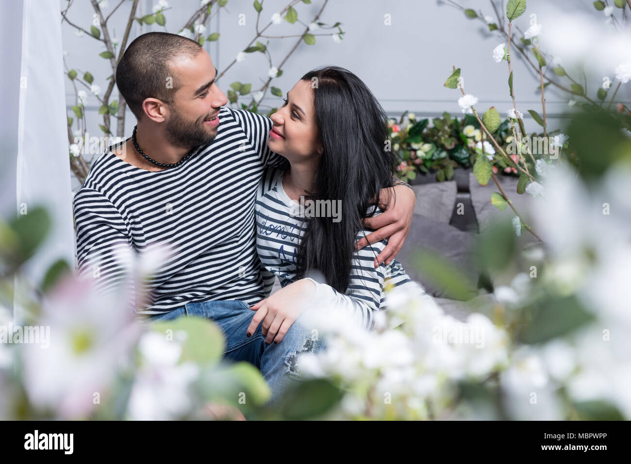 View through flowers on charming international couple in striped sweaters look at each other and hugging while sitting on the bed in the bedroom Stock Photo