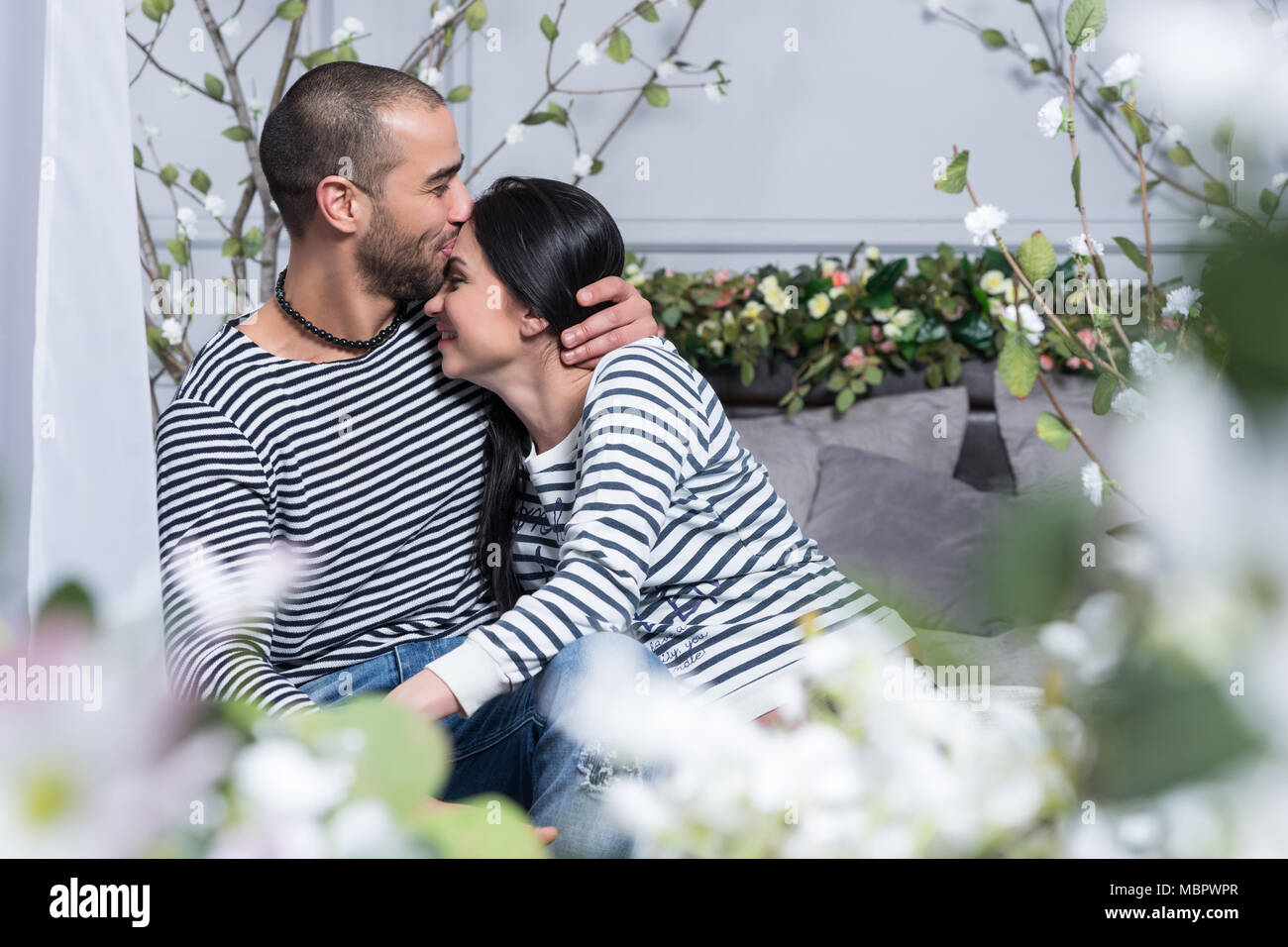 View through flowers on charming international couple in striped sweaters kissing and hugging while sitting on the bed in the bedroom Stock Photo