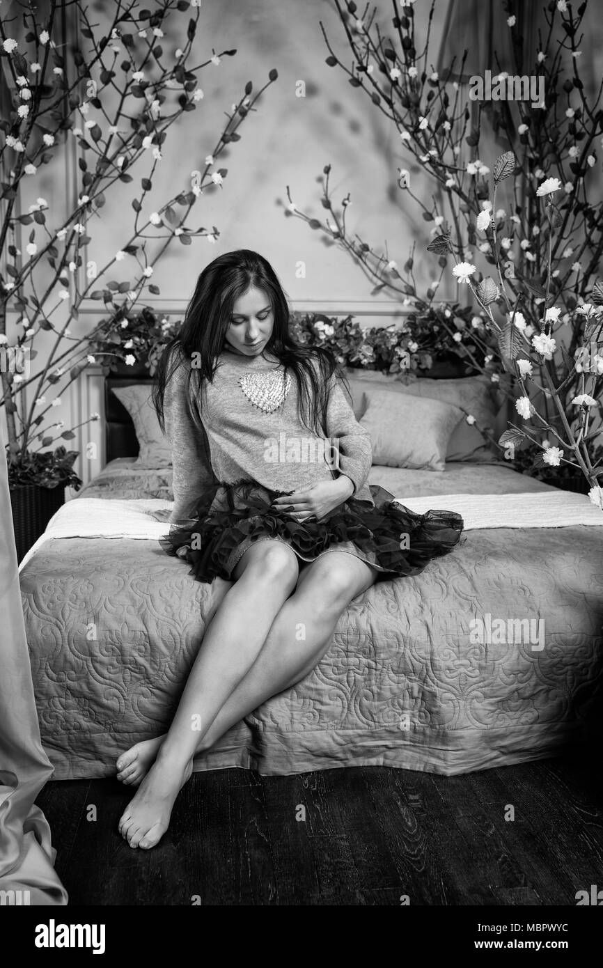 Black and white photo of cute pregnant brunette woman in a dress keeping hand on belly while sitting on the bed with flowers on bedstead in the bedroo Stock Photo