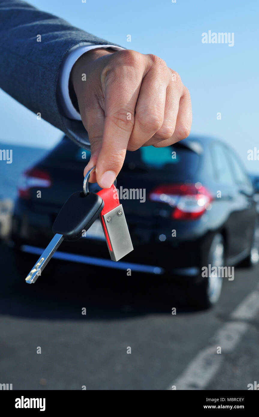 closeup of a young caucasian man, in a gray suit, offering a car key to the observer, with a car in the background Stock Photo