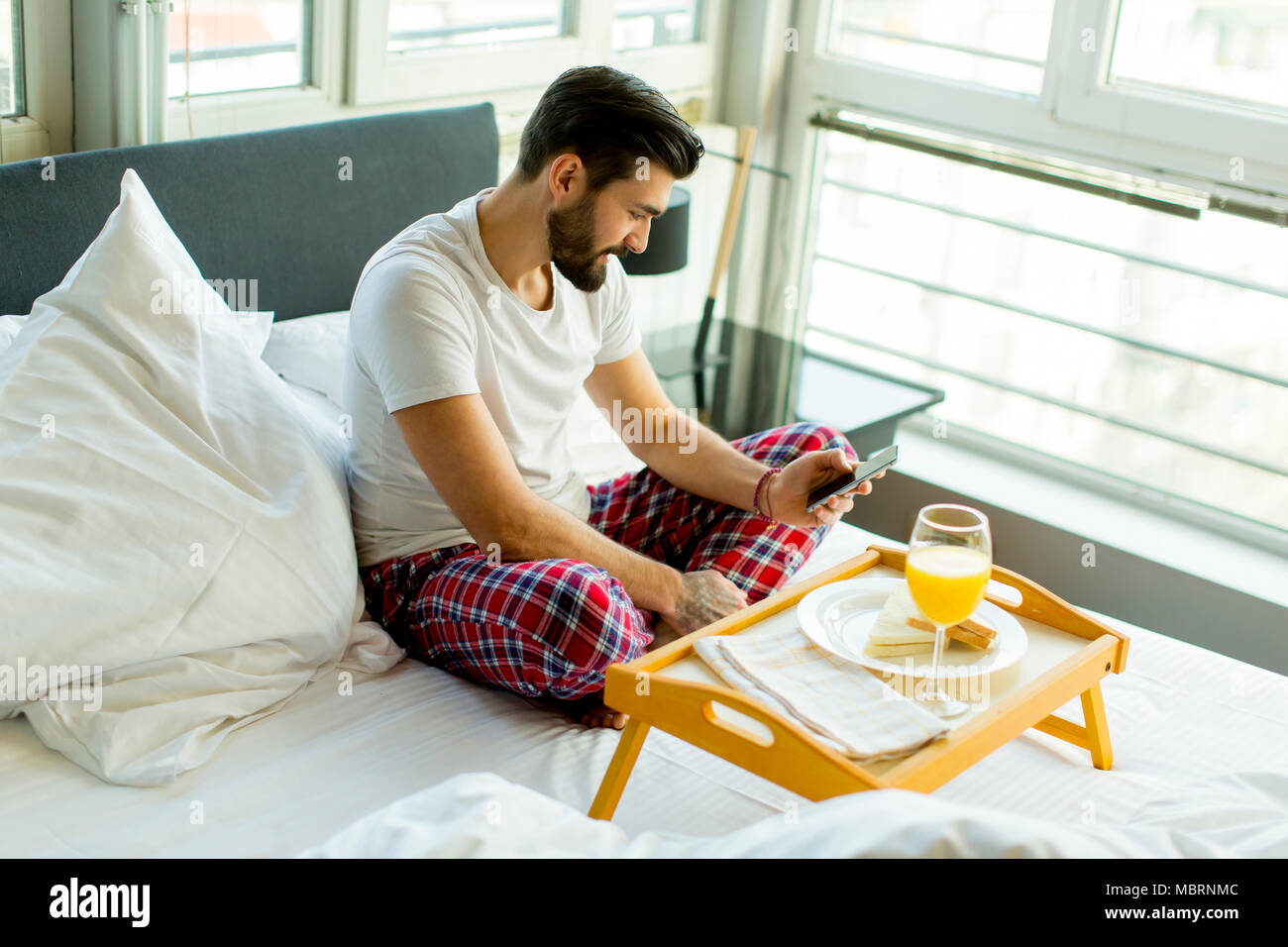 Young man eating breakfast in bed whilst using mobile phone Stock Photo