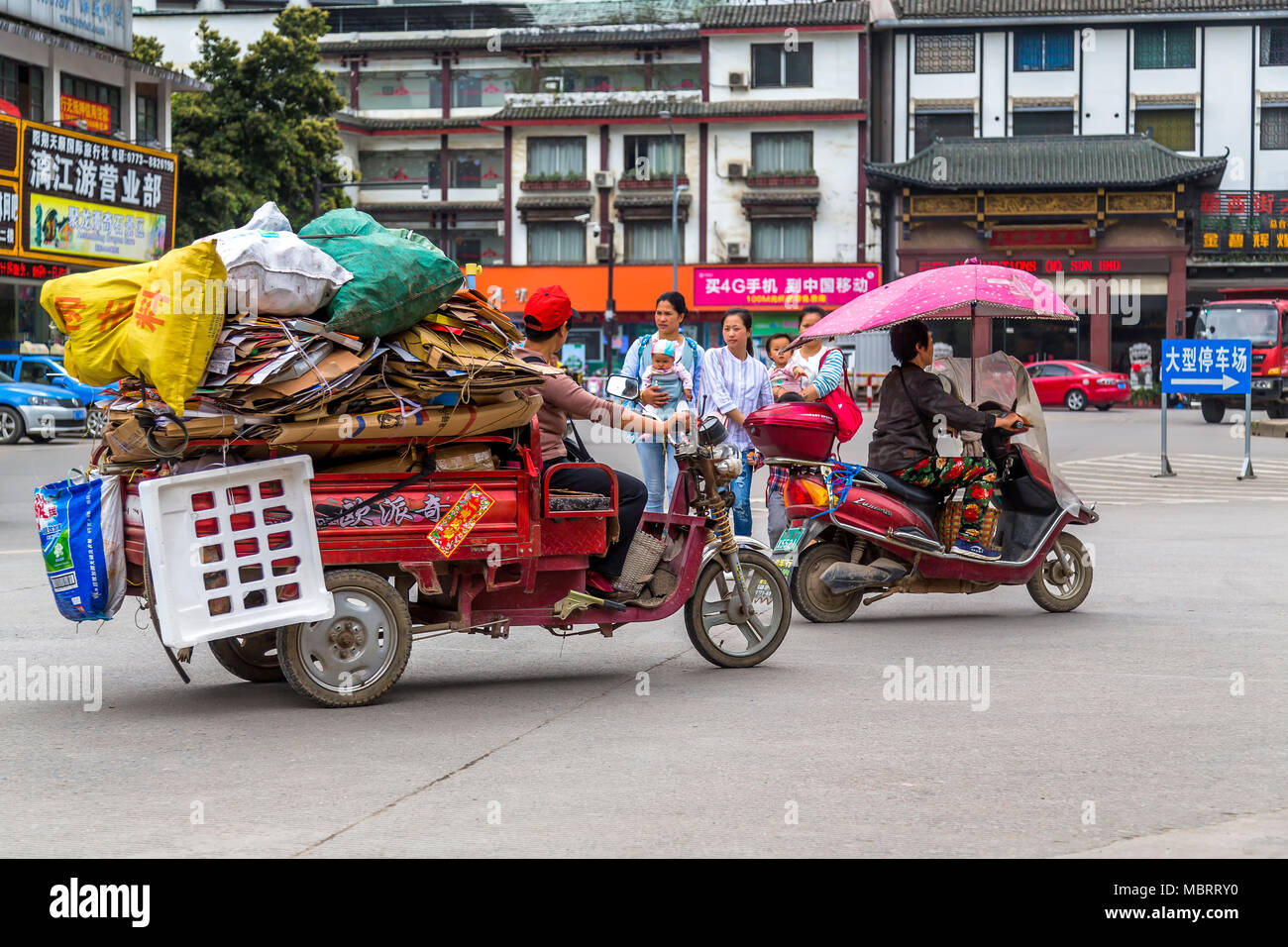Young mothers with their children cross a busy road in Yangshuo, China. A cardboard collector whizzes by on a 3 wheel motorcycle laden with waste card Stock Photo