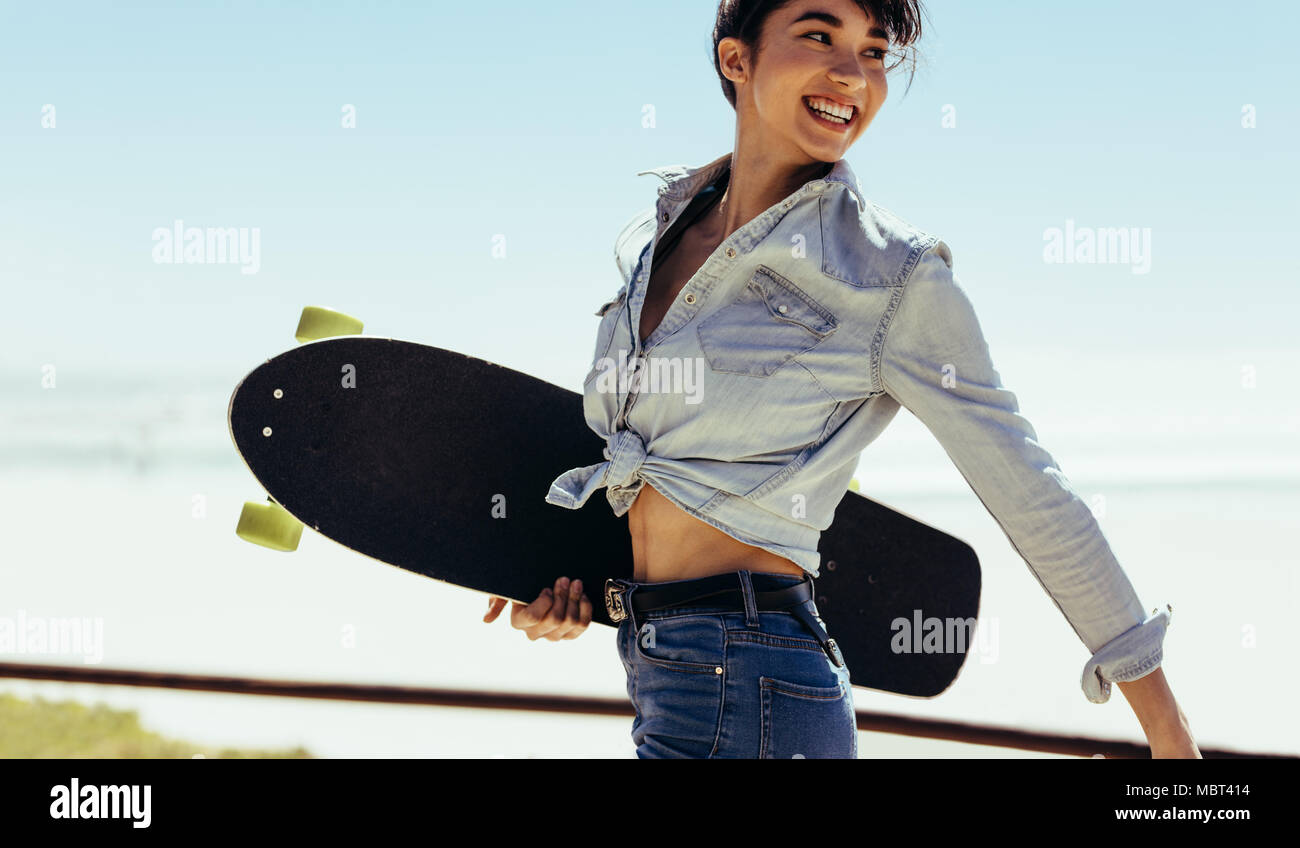 Woman walking outdoor on summer day with a skate board. Smiling caucasian female on a summer holiday. Stock Photo