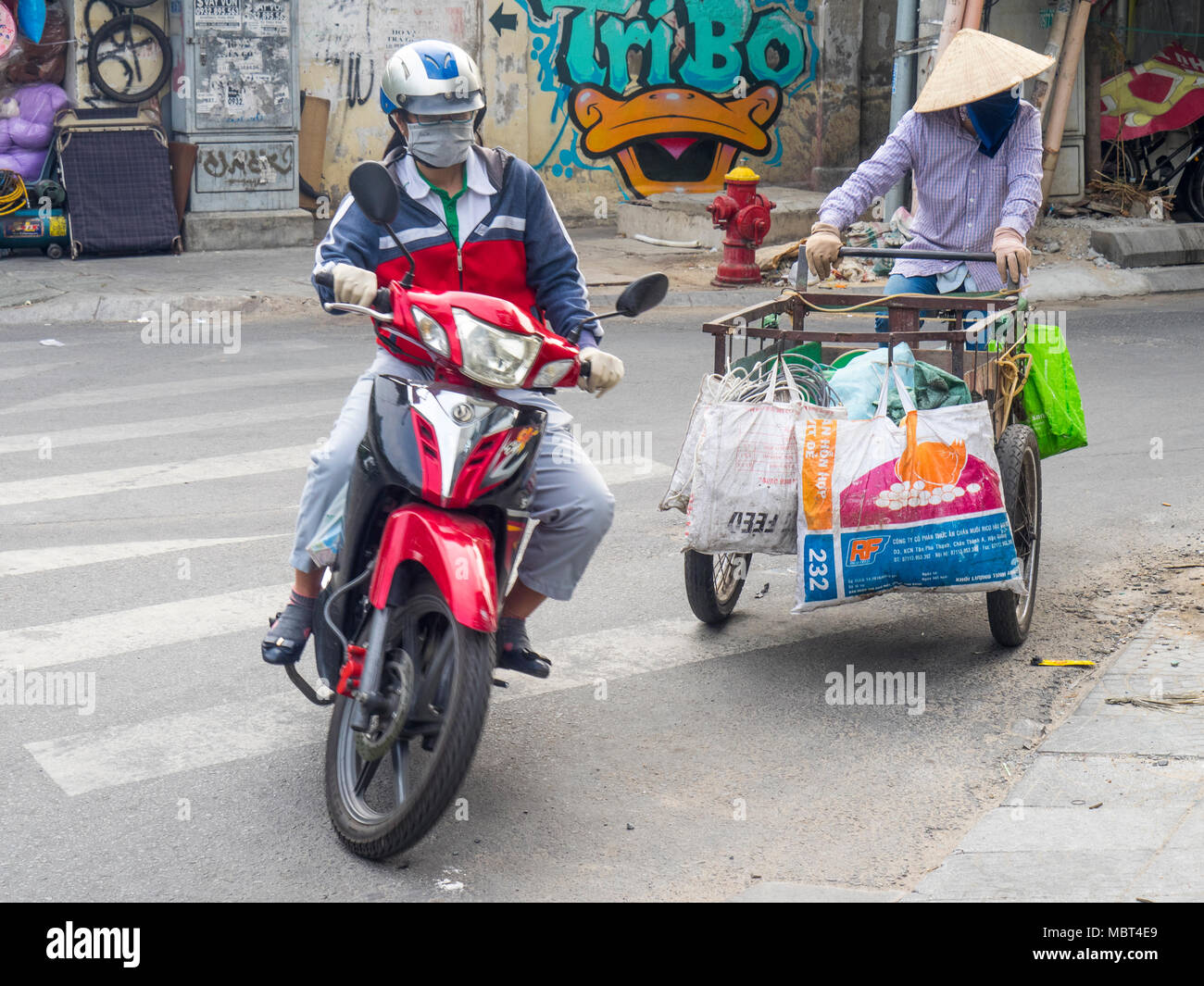 A  motorcyclist overtaking a Vietnamese woman wearing a conical hat riding a tricycle cargo bike in Ho Chi Minh City, Vietnam. Stock Photo