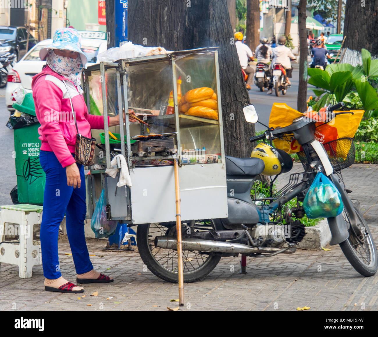 A female street vendor selling banh mi, bread rolls with fillings, from a cart attached to the back of a motorcycle in Ho Chi Minh City, Vietnam. Stock Photo