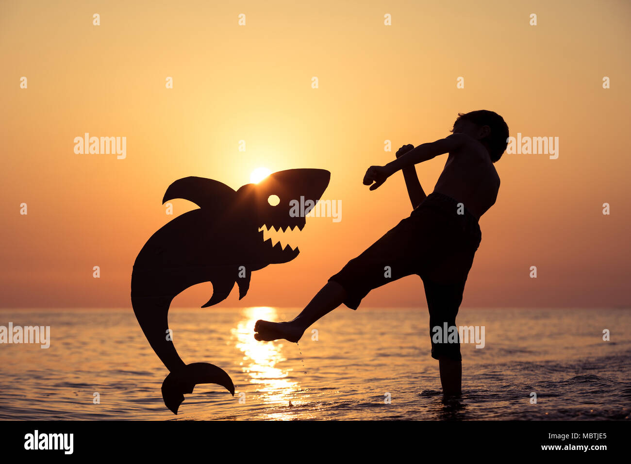 One happy little boy standing on the beach at the sunset time. He playing with a cardboard shark.  Kid having fun outdoors. Concept of summer vacation Stock Photo