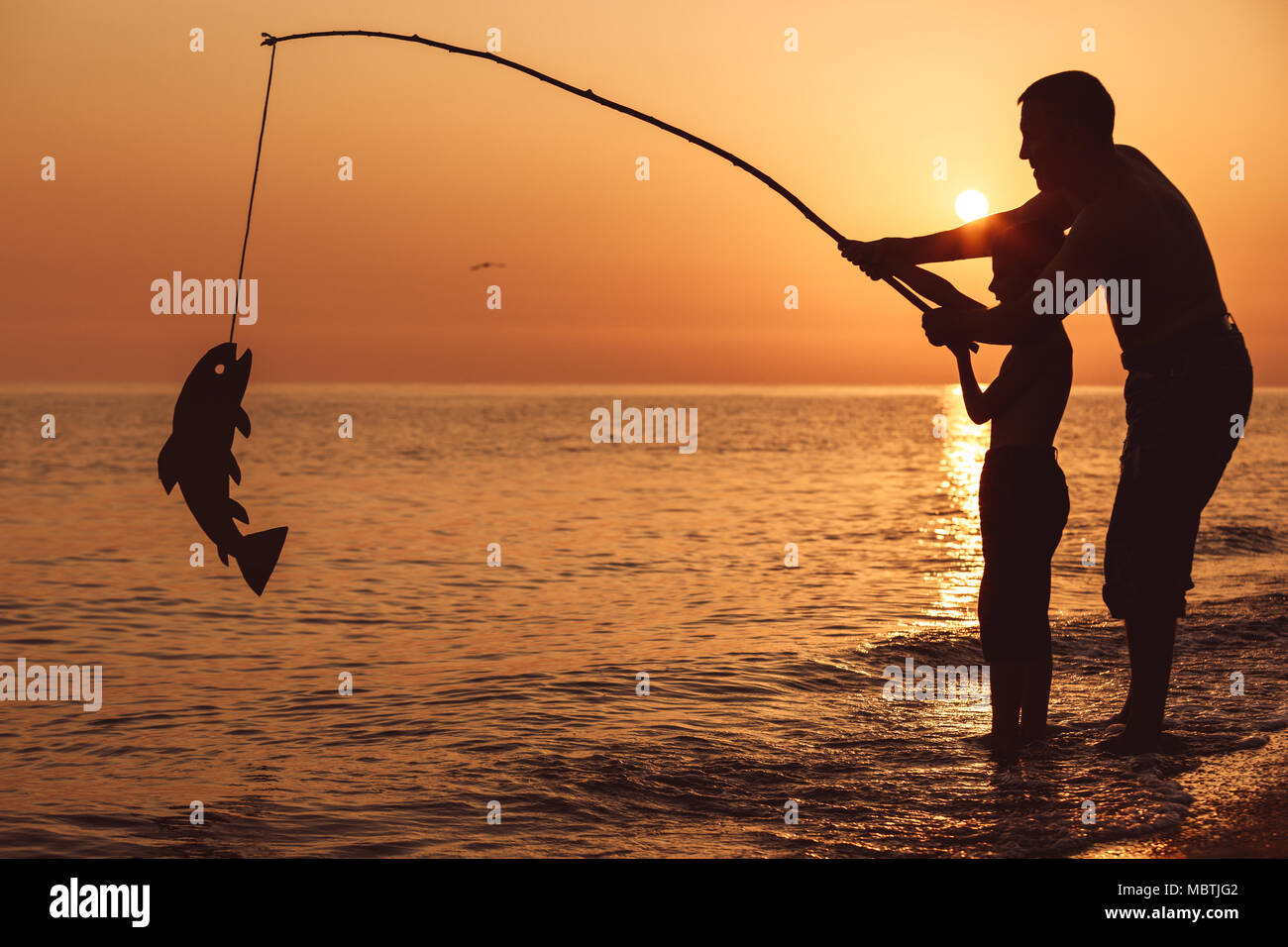 Father and son fishing on the beach at the sunset time. They playing with a cardboard fish. People having fun outdoors. Concept of summer vacation and Stock Photo