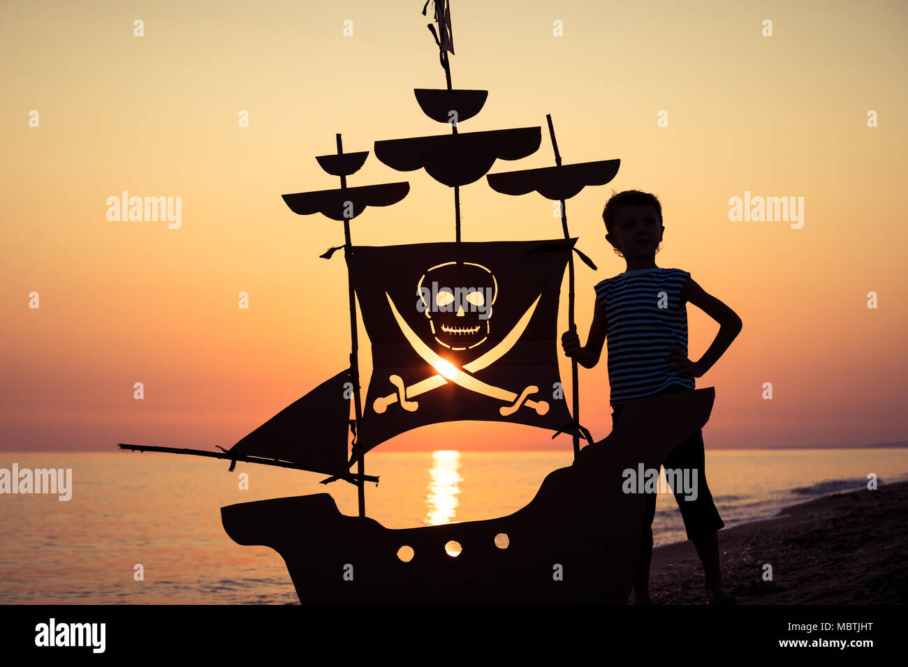 One happy little boy standing on the beach at the sunset time. He playing with a cardboard pirate ship.  Kid having fun outdoors. Concept of summer va Stock Photo
