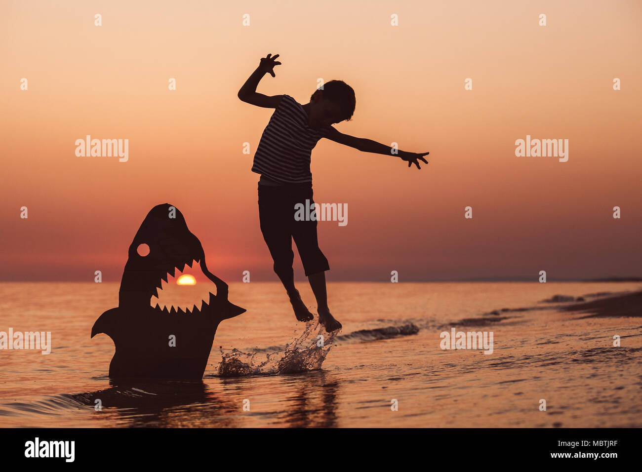 One happy little boy jumping on the beach at the sunset time. He playing with a cardboard shark.  Kid having fun outdoors. Concept of summer vacation. Stock Photo