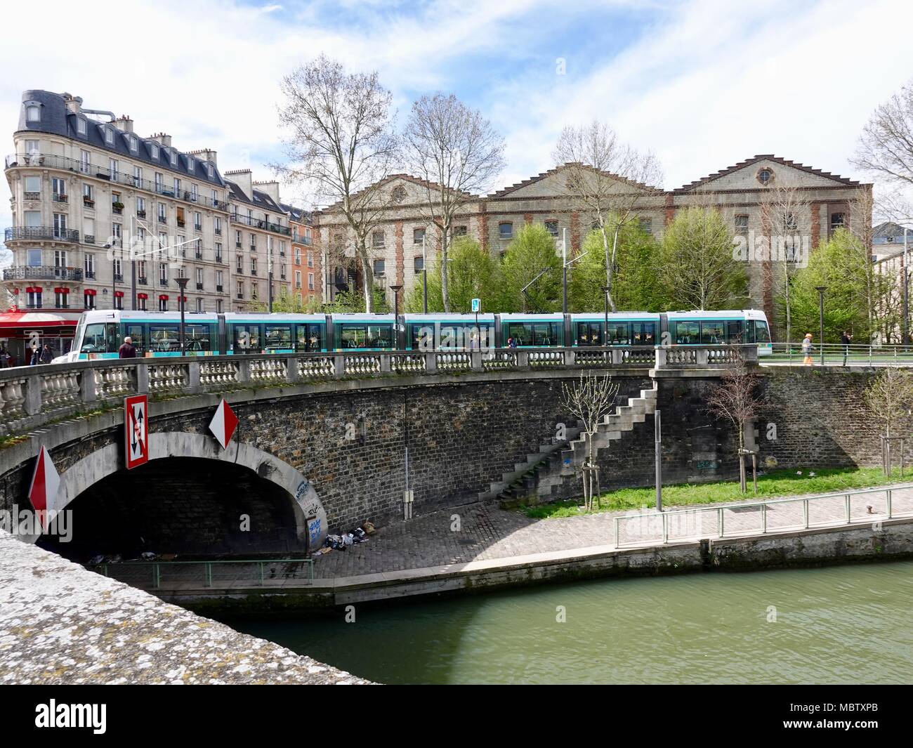 Tram with people, crossing over canal near Porte de la Villette with classic Parisian architecture in the background. Paris, France Stock Photo