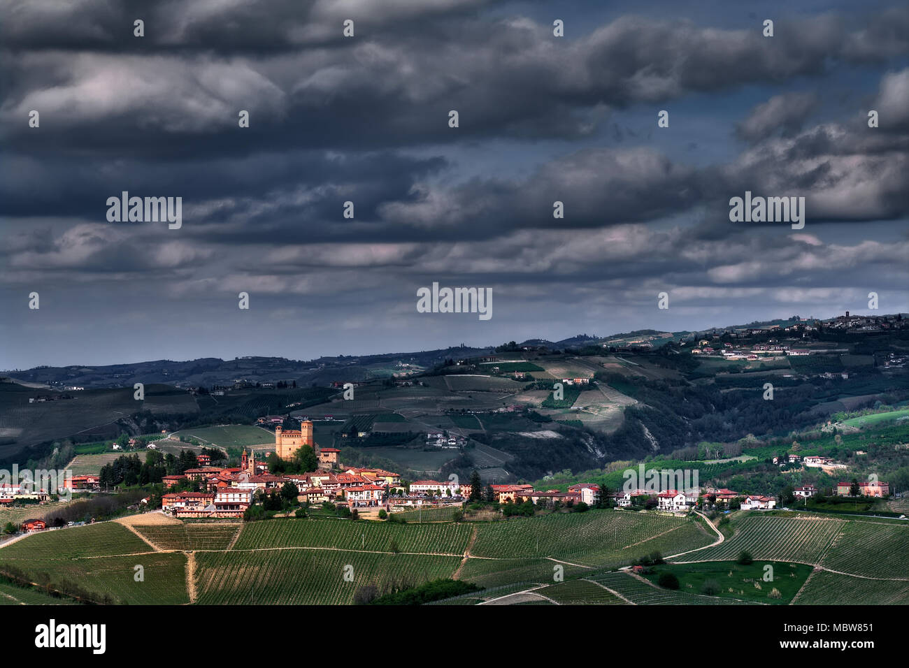 Wide view of the hills and vineyards of the Langhe, under a stormy sky, while a ray of sunshine illuminates Serralunga d'Alba and its towering castle. Stock Photo