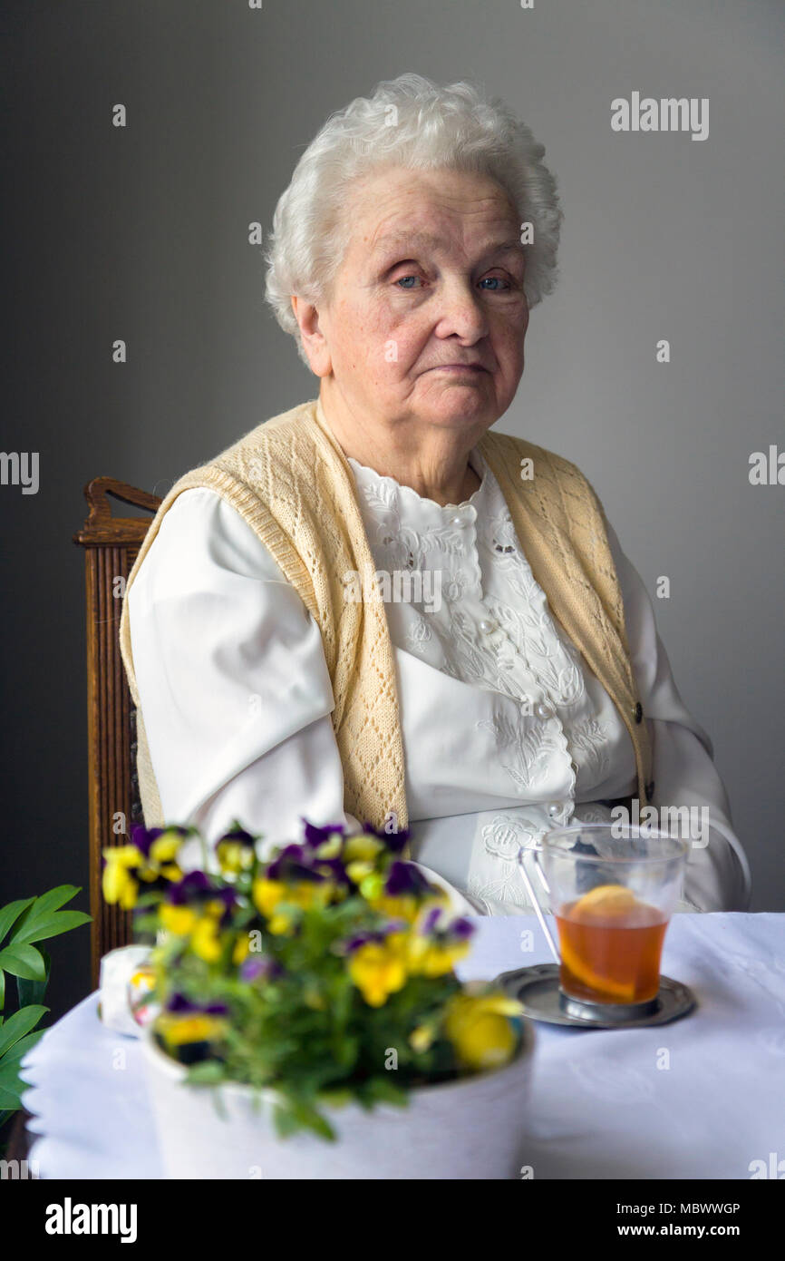 Old grey haired woman sitting at the table with flowers and a cup of tea Stock Photo