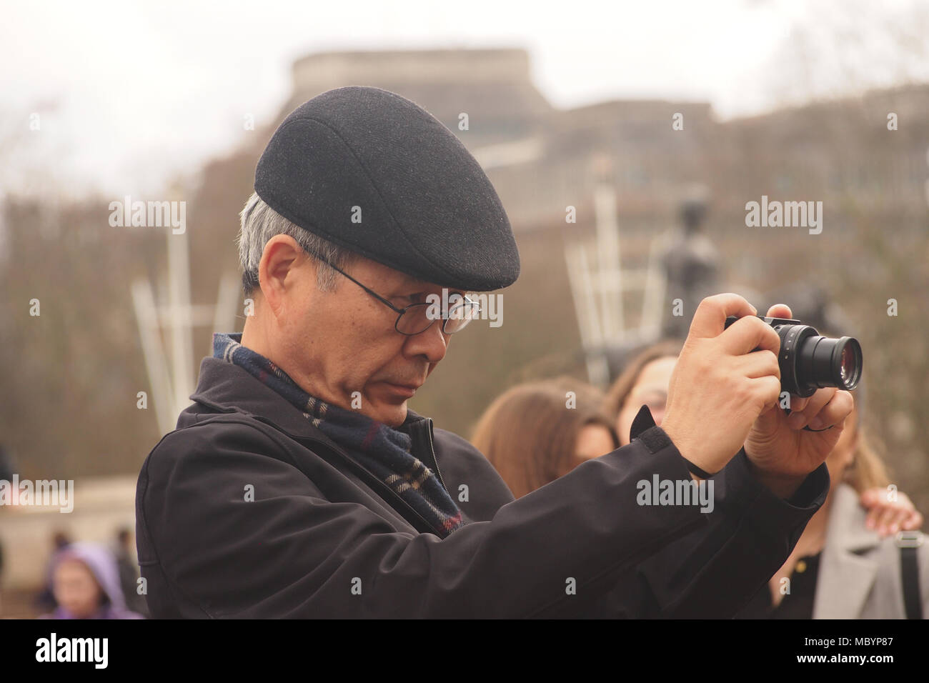 An older man taking a photograph with a camera in the area in front of Buckingham Palace, London, wearing a flat cap Stock Photo
