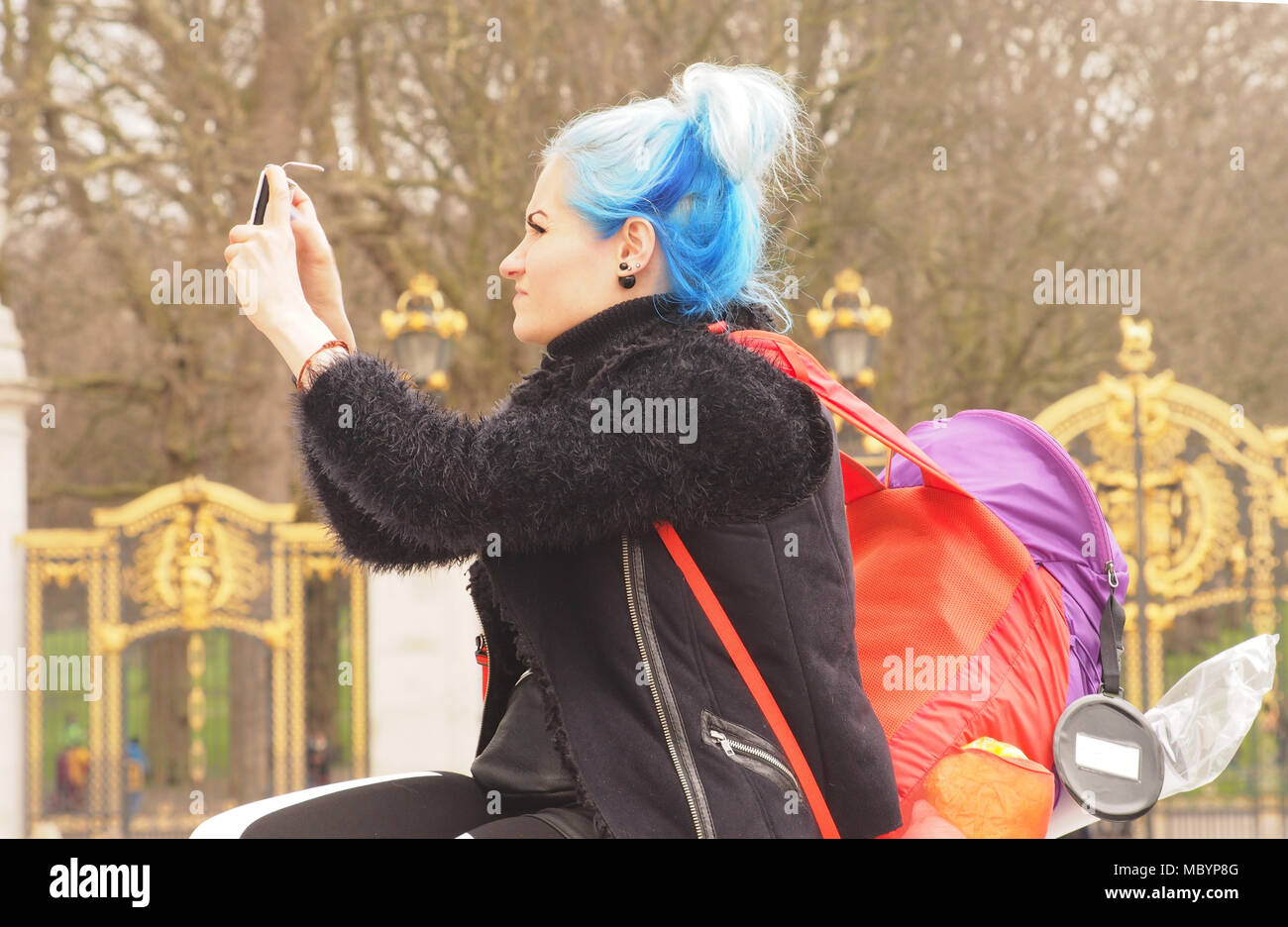 A young woman with striking blue hair taking a photograph on her smartphone with two brightly coloured backpacks in front of Buckingham Palace Stock Photo
