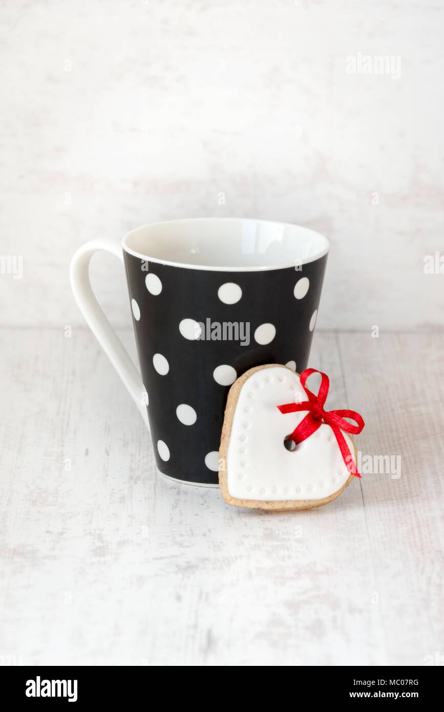 Black polka dotted coffee cup with a heart shaped homemade cookie over white wood background. Stock Photo