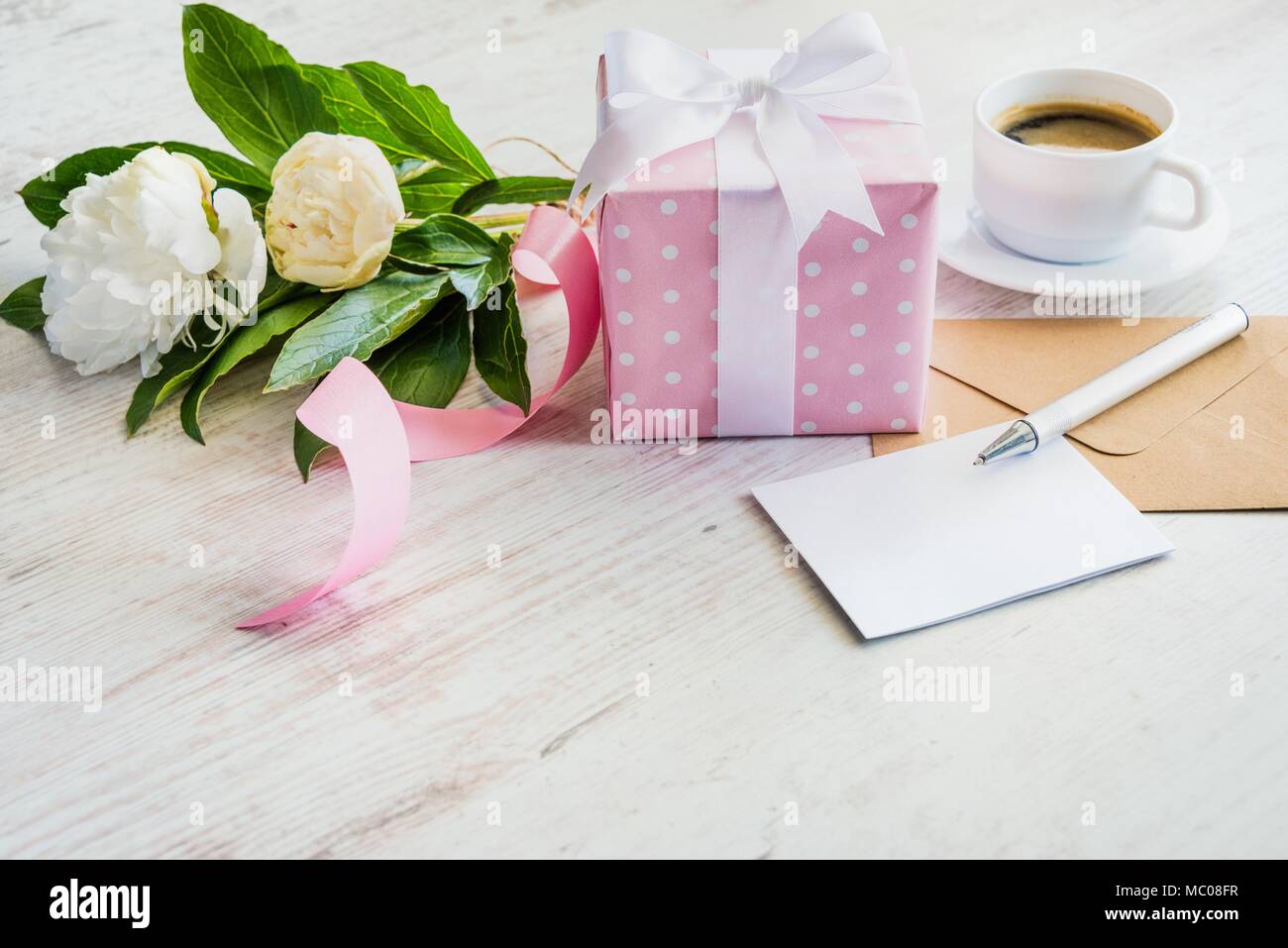 Pink dotted gift box, empty greeting card, kraft envelope, peonies bouquet and coffee cup over white wooden rustic table. Romantic holiday and event c Stock Photo