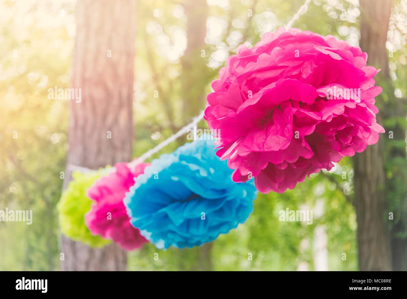 Close up of a colorful party garland made of paper flowers tied between trees in a park at an open air celebration event. Stock Photo