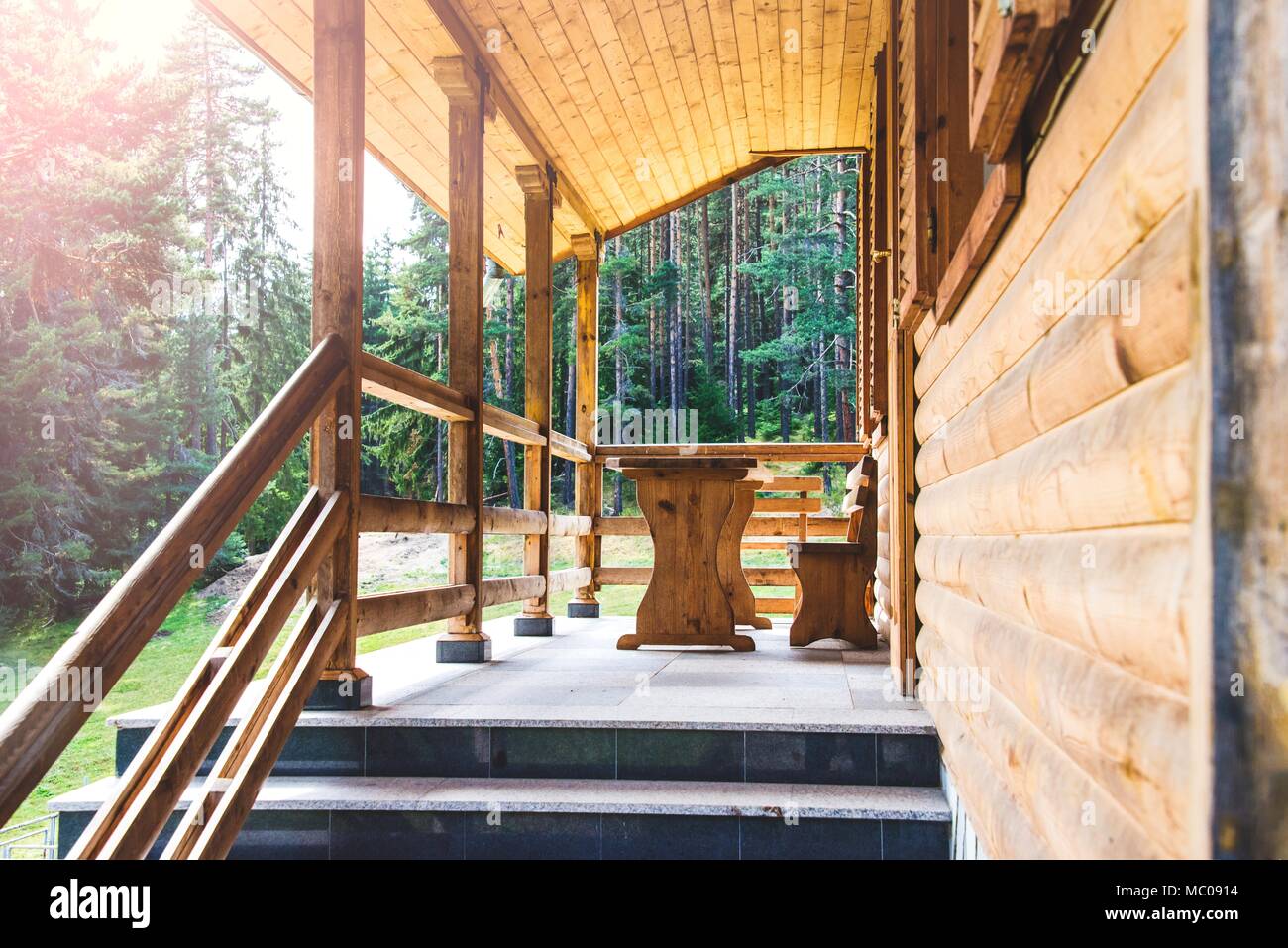 Side view of an open veranda in front of a wooden forest cottage. Pine forest under the sun rays in the background Stock Photo