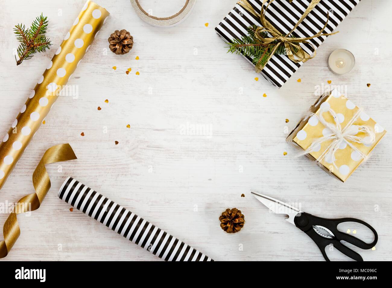 Gift boxes wrapped in black and white striped and goden dotted paper with, pine, cones, candle and wrapping materials on a white wood old background.  Stock Photo