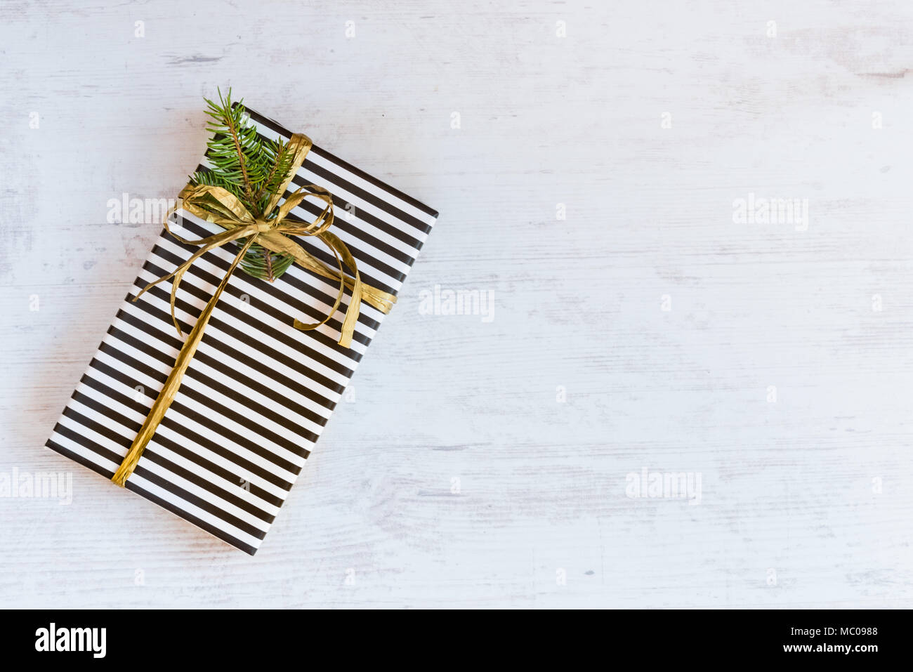 Gift box wrapped in black and white striped paper with golden ribbon and fir branch on a white wooden background. Christmas concept. Stock Photo