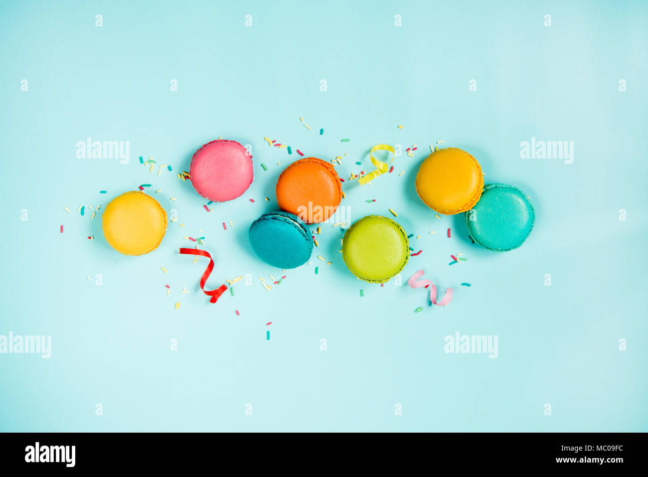 Top view of colorful macaroons, sugar sprinkles and party ribbons arranged over blue background. Stock Photo