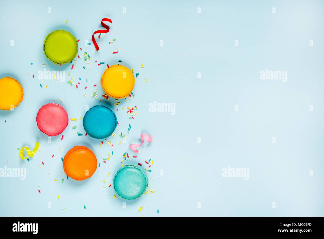 Top view of colorful macaroons, sugar sprinkles and party ribbons arranged over blue background. Copy space. Stock Photo