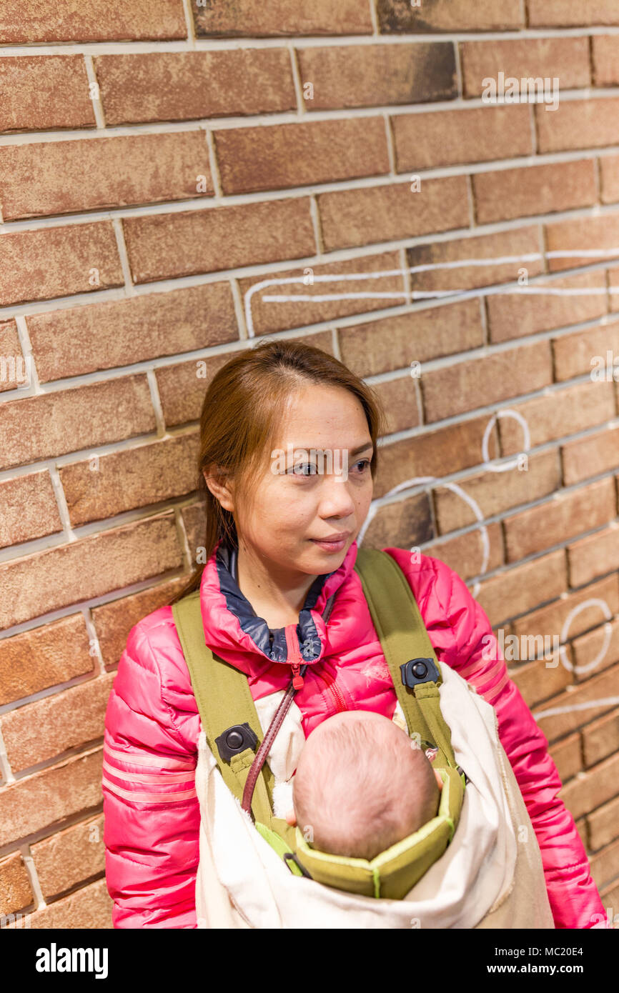 Mother carrying baby in baby carrier standing next to brick wall. Stock Photo