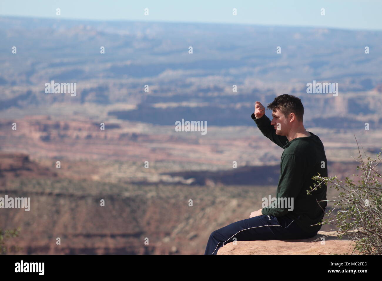 A man with a green long sleeve shirt shading his eyes from the sun while sitting on the edge of a canyon overlooking Canyonlands National Park, Utah Stock Photo