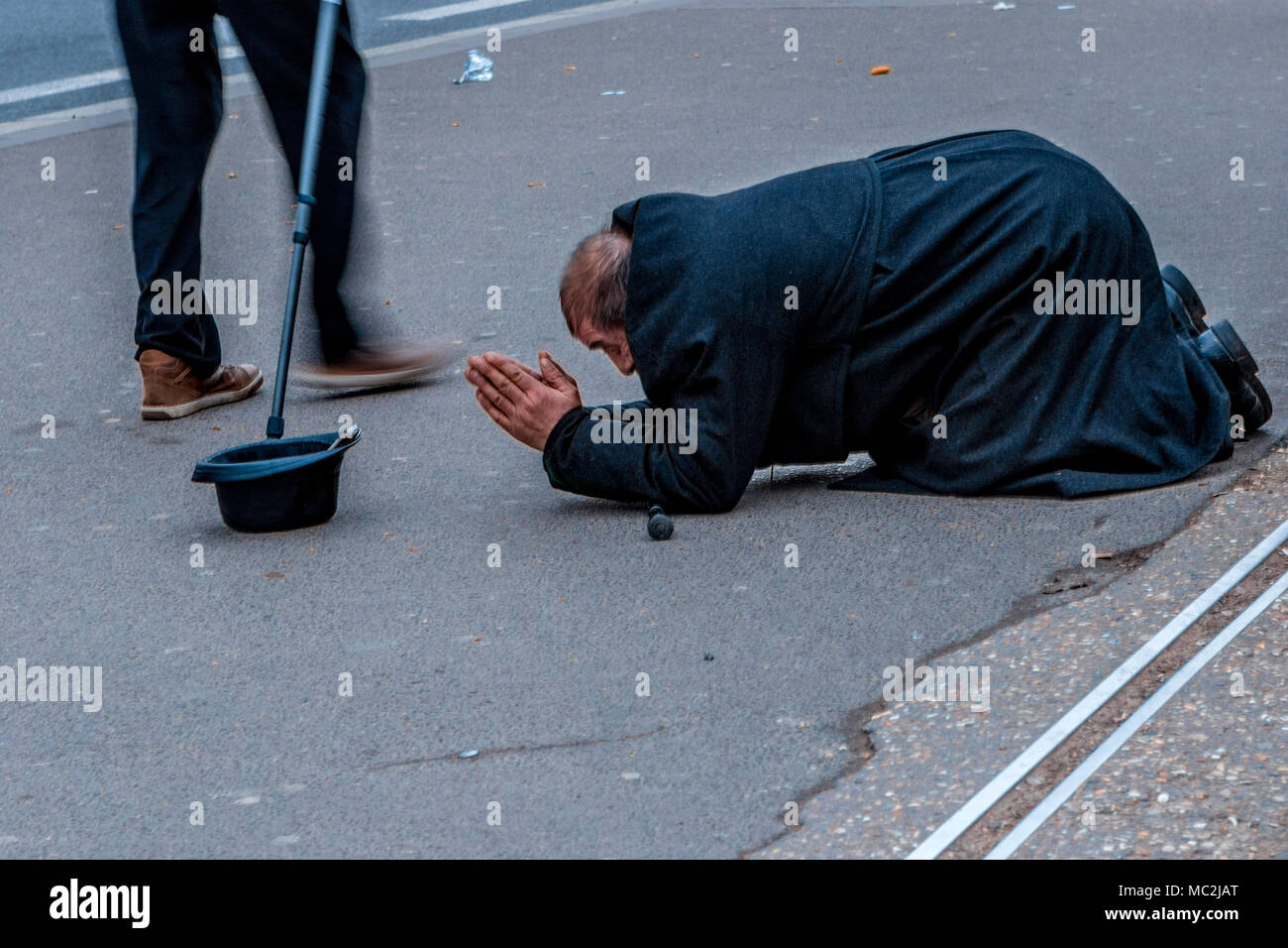 Prostrate beggar with big coat in attitude of prayer with begging hat; a passerby with walking stick walks by this eastern man Stock Photo