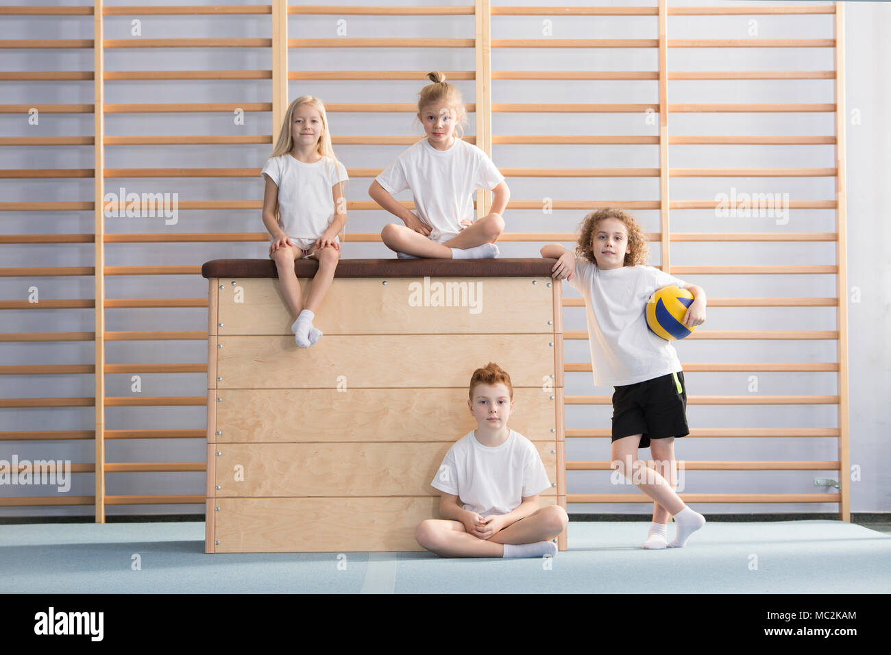 Boy with a ball next to a wooden vaulting box on which smiling children are sitting in the gym hall Stock Photo