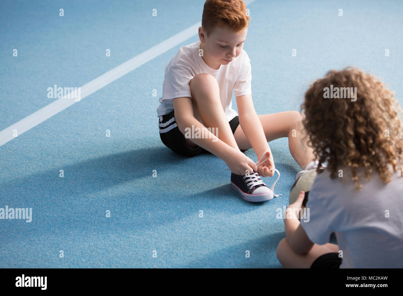 Young boy sitting on a blue floor and tying the shoelaces before physical education classes Stock Photo