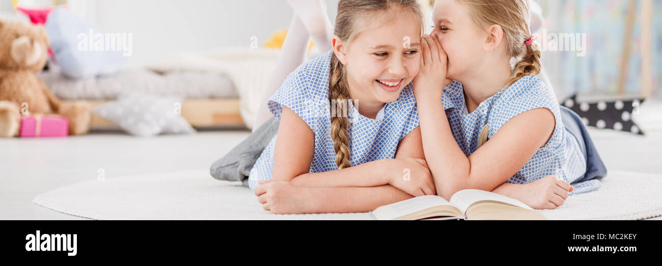 Happy young girl whispering a funny joke to her sister while spending time together in a bright kid's bedroom Stock Photo