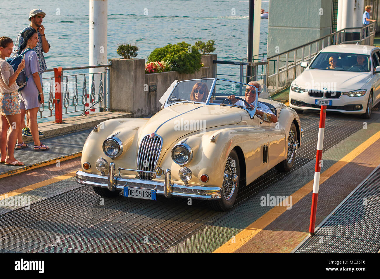Happy man drives out his vintage beige Jaguar XK120, a sports car manufactured by Jaguar circa 1950, from the ferry boat at lake Como Stock Photo