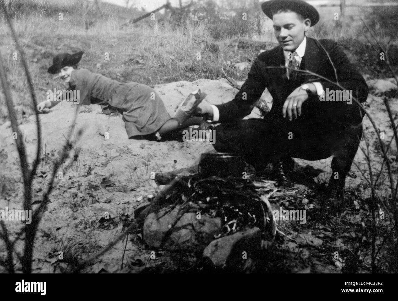 A young man cooks over a campfire while his date watches, ca. 1920. Stock Photo