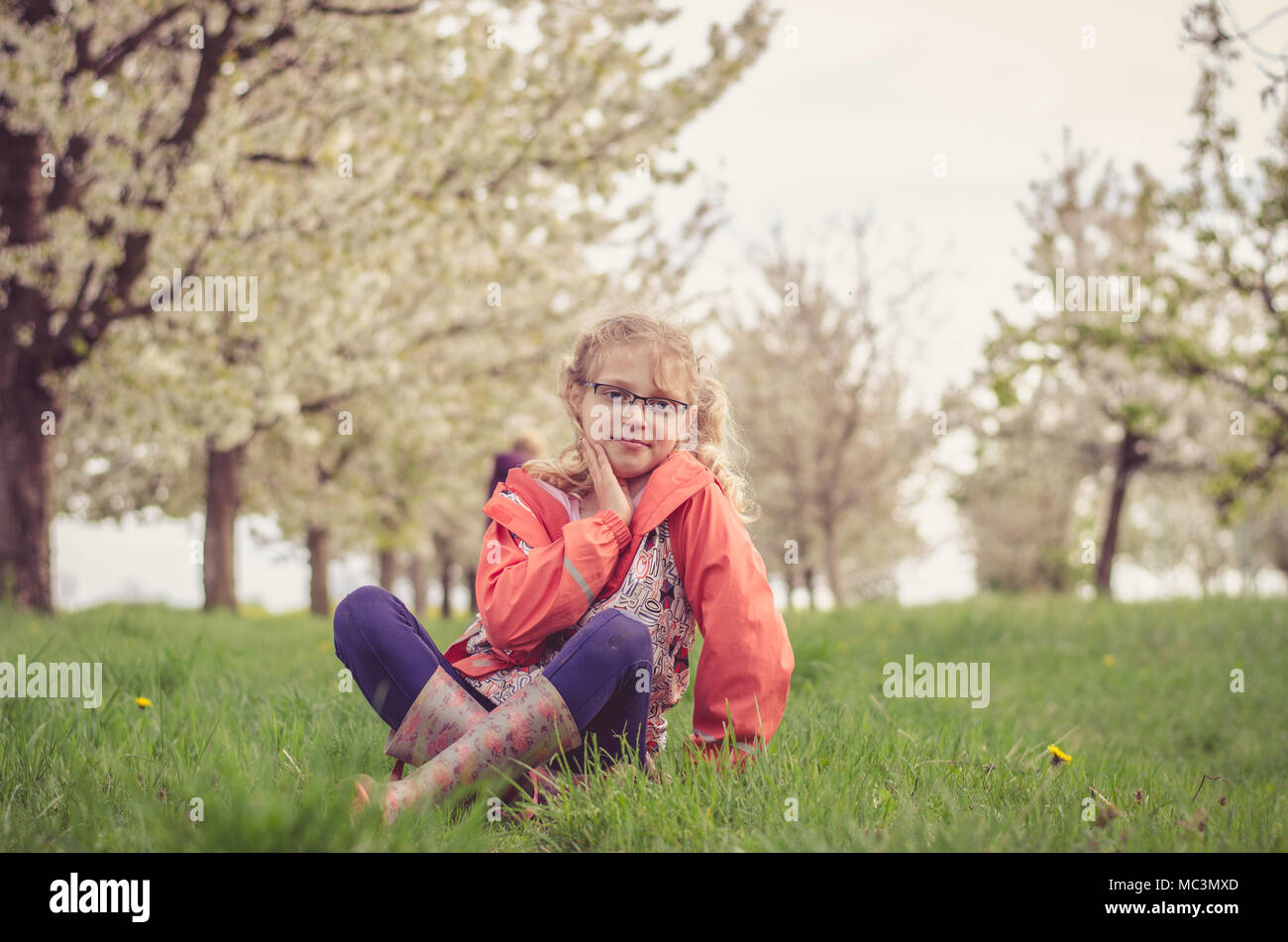 adorable blond girl sitting in green grass under trees with blossoming white flowers in spring season Stock Photo