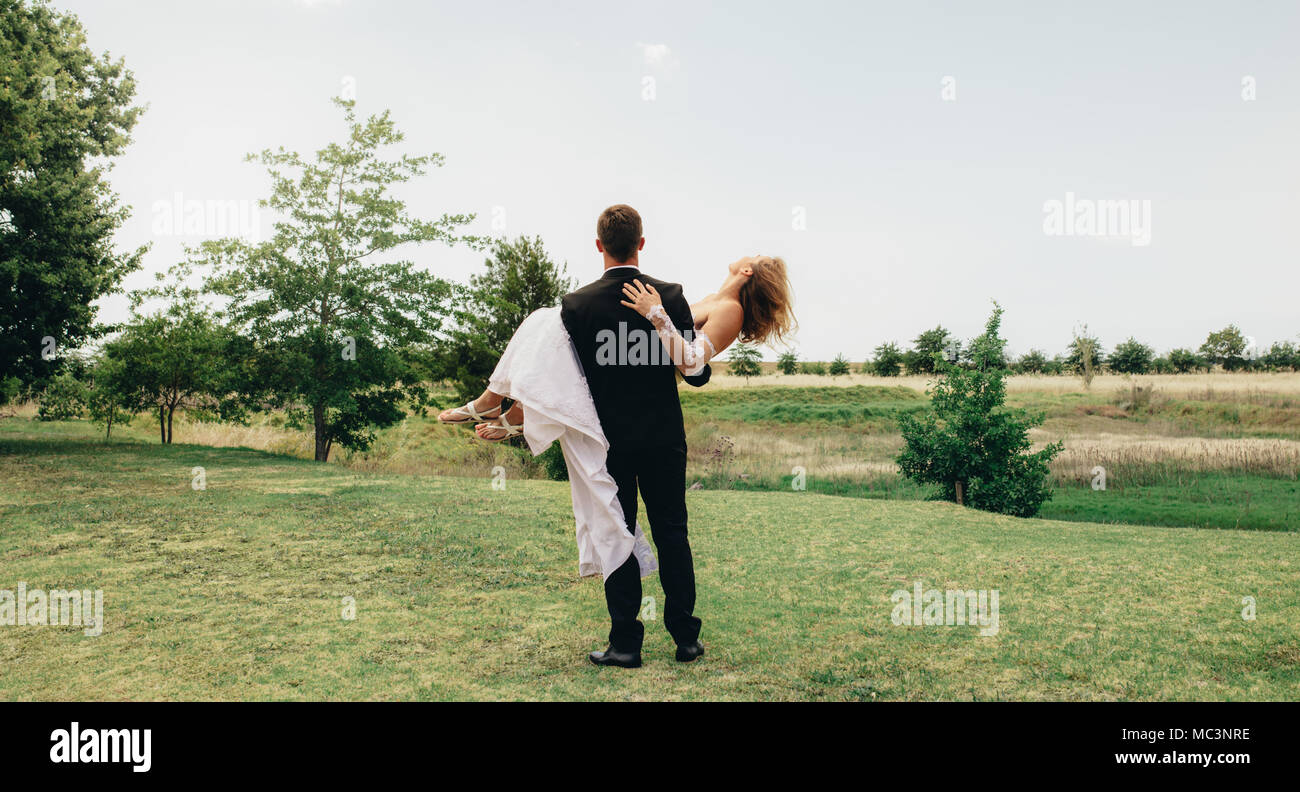 Bridegroom carrying his bride in his arms and standing outside. Rear view of newly wed couple celebrating their marriage outside. Stock Photo