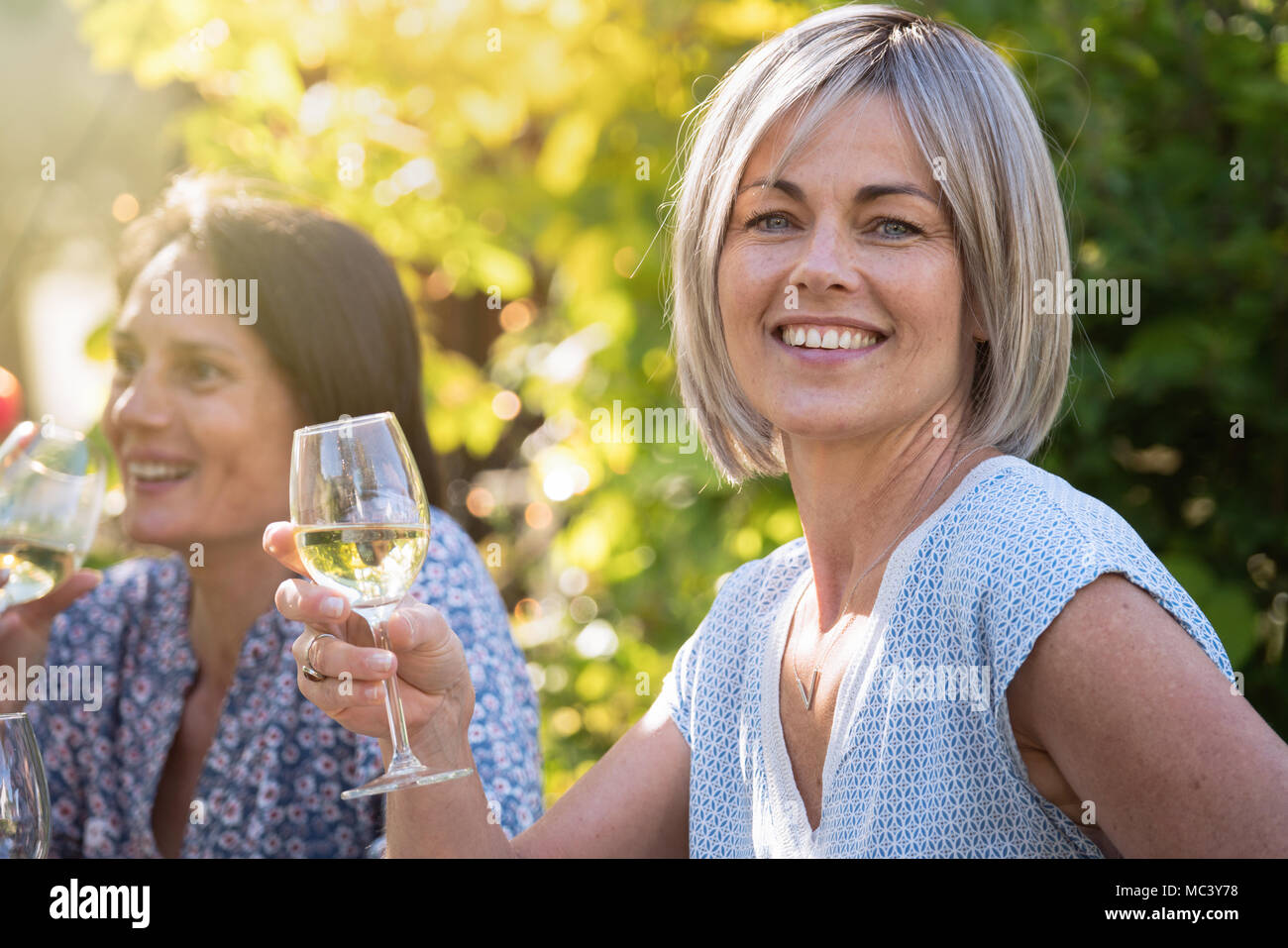 Group of friends gathered around a table in a garden to have a good time together. Focus on a beautiful woman looking at camera a drink at hand Stock Photo