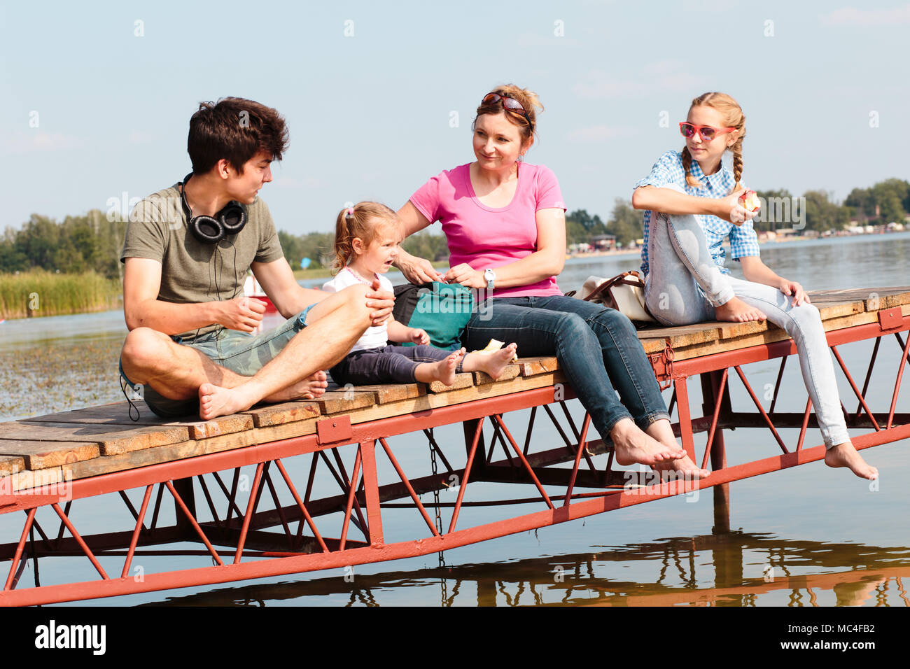 Family spending vacation time together having a snack sitting on jetty over the lake on sunny day in the summertime. Quality family time at weekend Stock Photo