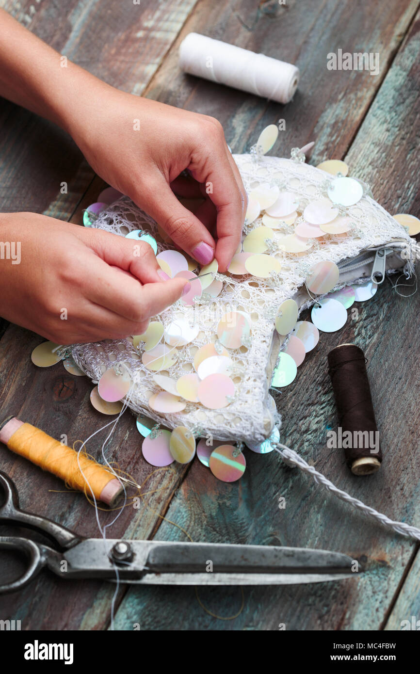 Close shot of female hands decorating purse with sequins using thread needle and scissors following diy ideas on wooden table Stock Photo