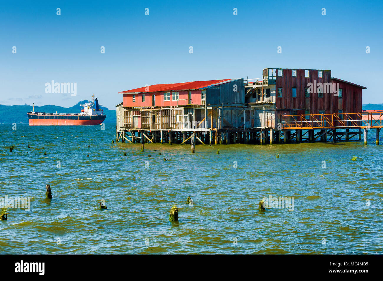 An aabandoned fish cannery along with a distant freighter anchored in the river is sceen from Astoria's waterfront along the Columbia River in Oregon. Stock Photo